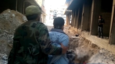 A militant or soldier holding a blindfolded man in front of a dirt pit between buildings
