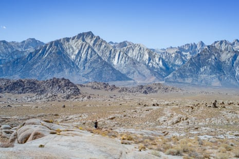 A lone hiker walks across the Alabama Hills beneath Mount Whitney's jagged, snow-streaked peaks