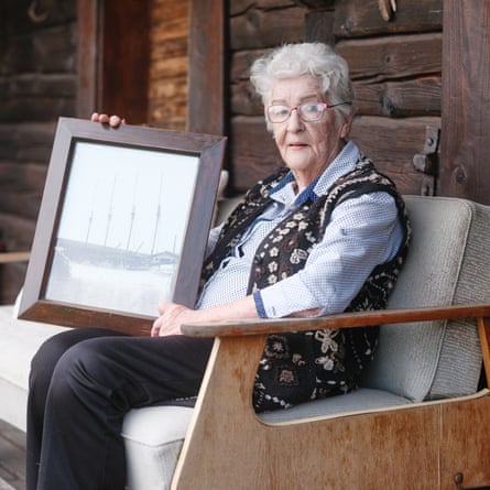 Aasa Sarnik, an elderly woman with white hair, sitting on a couch outside, wearing a blue shirt and embroidered brown sleeveless coat, holding a large black and white picture of a ship