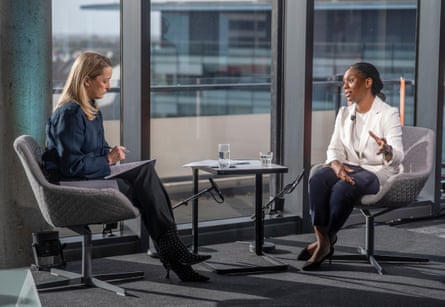 Laura Kuenssberg and Kemi Badenoch sit in grey swivel chairs either side of a small table in front of a large window. Kuenssberg is looking down at her lap, where she has her notes, as Badenoch speaks.