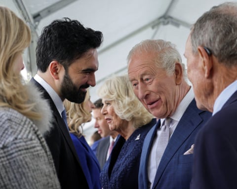 A man in a suit smiles as another man in a suit speaks, among a group of other men in suits and women in professional wear.