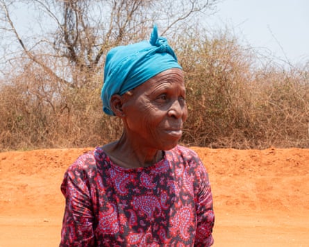 An African woman wearing a blue headwrap and floral dress against a background of red earth and trees.