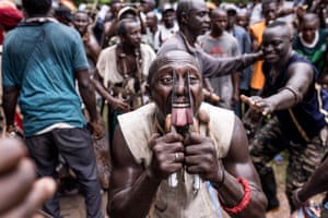 A man holds two large knives to his tongue as other men dance and gather behind him