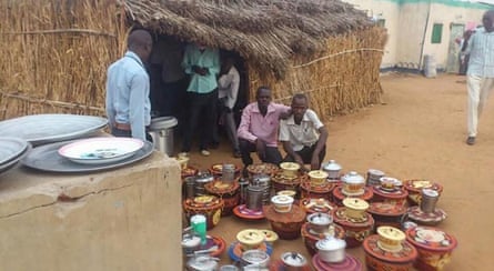 Two men sit on the ground outside a straw hut, one with his arm around the other. Another man looks on. Lots of baskets with cooking pots on them are lined up on the ground in front of them