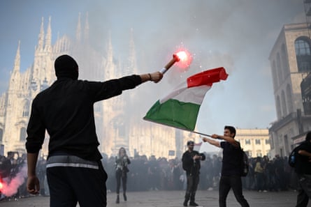 A masked man holds a lit flare while another waves an Italian flag during a protest