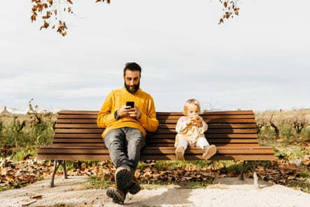 Posed by model Father and daughter sitting on a bench in the park in autumn, father using smartphone, daughter eating an apple