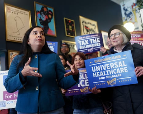 a woman speaks in front of people holding signs that read 'tax the billionaires' and 'universal healthcare'