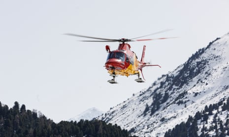 A helicopter flies above a treeline with snowy mountains in the background