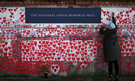 A person writes on the national Covid memorial wall – she is standing on tiptoe to paint a small red heart on the white wall to join those already depicted, which have names of those who died from Covid written within them