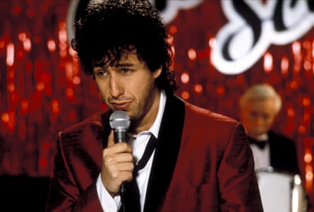 A man with dark curly hair, red glittery jacket, white shirt and black tie, holding a microphone. The background is very glittery