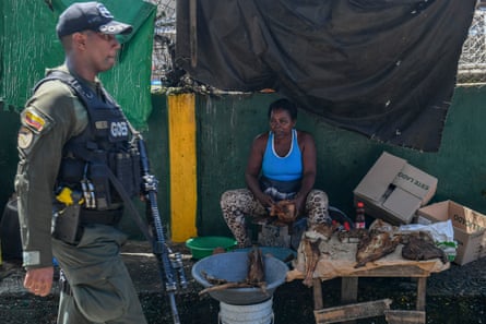 An armed soldier walks past a woman sitting under a tarp next to a table of cooked fish.