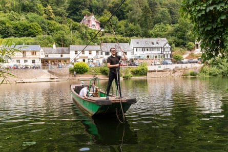 A man standing on the front of a tiny ferry, pulling it across a river, with two people and a dog in, and buildings on the far bank