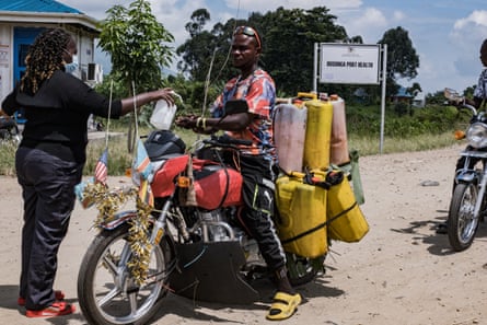 A woman dressed in black and wearing a blue face mask squirts hand sanitiser on a man riding a motorcycle, which is heavily laden with what appear to be plastic fuel cans.