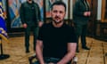 Volodymyr Zelenskiy, wearing dark green trousers and a black T-shirt, sits in a chair and looks at the camera. He has a serious expression and it holding his hands in his lap.