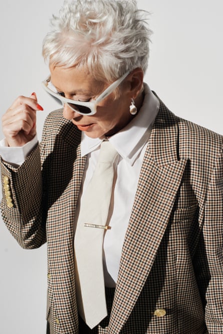 Denise Welch pictured from the waist up, wearing a pale-brown trouser suit, a white shirt and tie with a tie-pin, and black, white-rimmed sunglasses, looking down to her right with her right arm bent at the elbow , against a pale-grey background