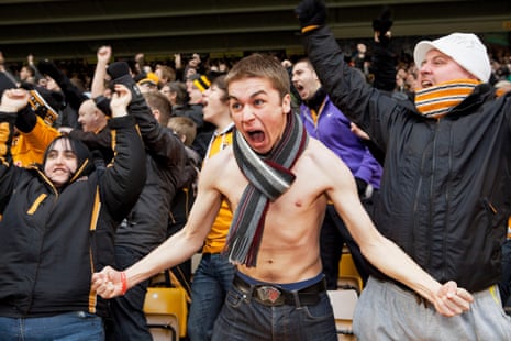A young man with no shirt and a scarf round his neck shouts with joy while surrounded by other fans in jackets and hats