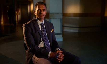 Siva Anandaciva: he is seated in a darkened interior which looks like a hospital corridor, with light shining behind him and wears a dark jacket, white shirt and dark blue tie with coloured spots on it.