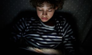 Young boy on sofa in dark room watching touchpad illuminated with screen light