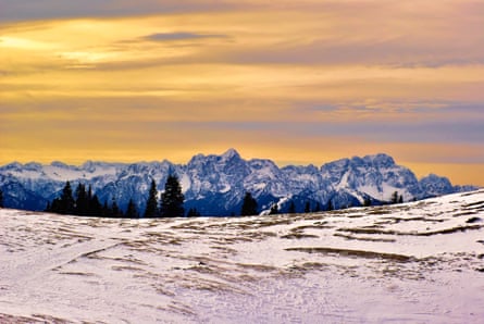 A yellow sunrise on a plateau dusted with snow, with the tops of mountains and fir trees in the background