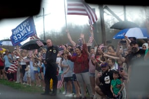 People holding US flags and a Trump/Vance sign line the side of a road