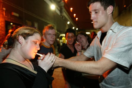 A man in a white shirt holds a lighter out to light the cigarette of a woman in a dark top