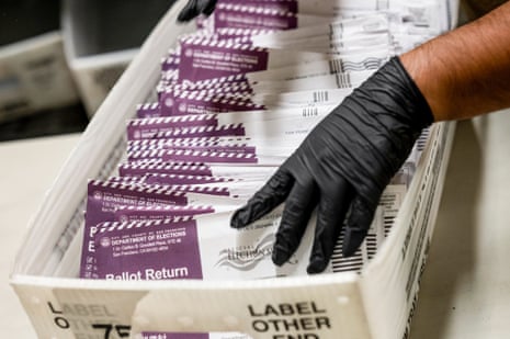 An election worker's gloved hand sorts purple "Ballot Return" envelopes in a tray