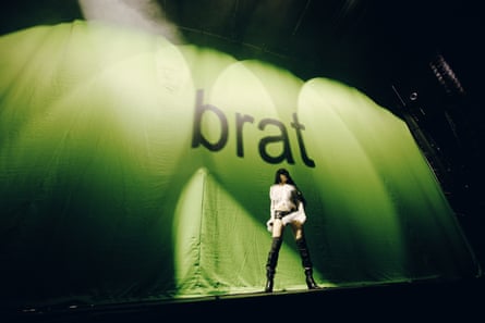 A tiny view of a woman with dark hair and white top and high thigh length books in front of an outsided green backdrop reading ‘brat’