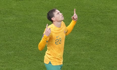 A Socceroos player in a yellow jersey and green shorts points his index fingers up to the sky and looks up after scoring a goal