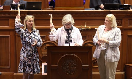 Three white women with blond hair hold small models of spines, standing behind wooden lectern in front of raised wooden dais, as if in a legislative chamber.
