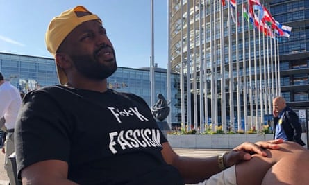Magid sits outside the European parliament. Flags of EU countries are in the background. He is wearing a black t-shirt that says: F**k fascism
