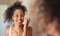 Young black woman applying skin cream<br>Young black woman applying moisturising cream to her skin while standing in front of the mirror in the bathroom. African american girl applying face cream while smiling. Beauty hydrating moisturizer and skincare routine concept.