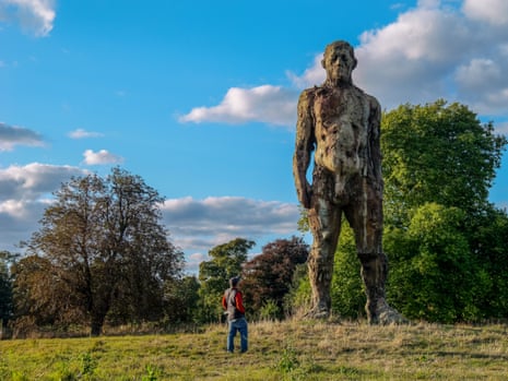 A person stands in a field looking up at a towering weathered sculpture of a human figure