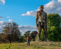 A person stands in a field looking up at a towering weathered sculpture of a human figure