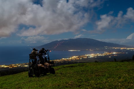 Hunters on a slope with city lights on the shore of the ocean and a large mountain in the background.
