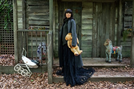 A white woman with very strikingly sculpted dark hair wears a long dark dress with a grey jacket and holds a teddy bear in front of a cabin and some vintage toys