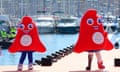 The Olympic and Paralympic mascots, like red Phrygian caps on legs, pose on a quayside, with  yachts in the background.