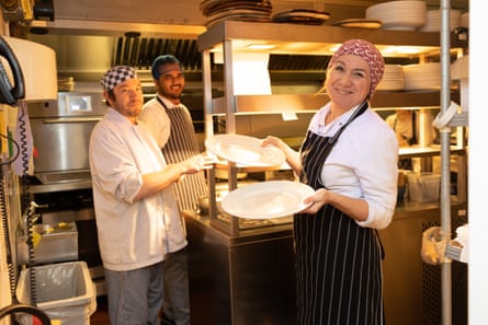 A smiling woman in an apron stands holding plates in a restaurant kitchen while two other smiling catering workers look on
