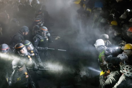 Cops in riot gear face off with students, some of them wearing hard hats and bicycle helmets, amid a cloud of black smoke