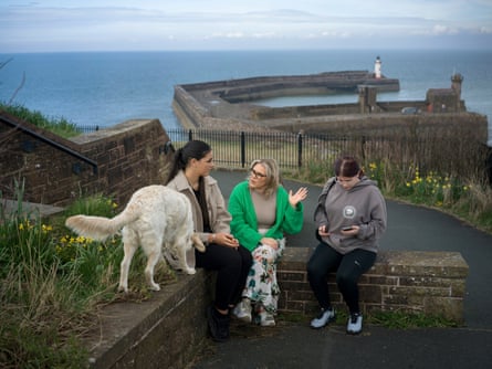 Three women sit on a wall with a dog, with a view of the sea and a lighthouse behind them.