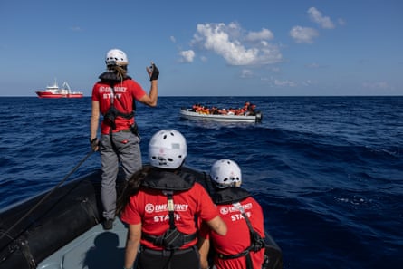 Three people wearing red t-shorts and white helmets in teh front of a boat sailing towards a small crowded small boat with an outboard motor.