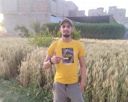 A young Arab man in a T-shirt and baseball cap poses by a wheat field, making a gun-like gesture