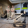 Capt Liam Kidman and Maj Andrew Boyce stand arms crossed beside a bomb-disposal robot and truck