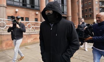 Adam Carruthers  in black hoodie, black mask and sunglasses walks along a street with photographers taking his picture