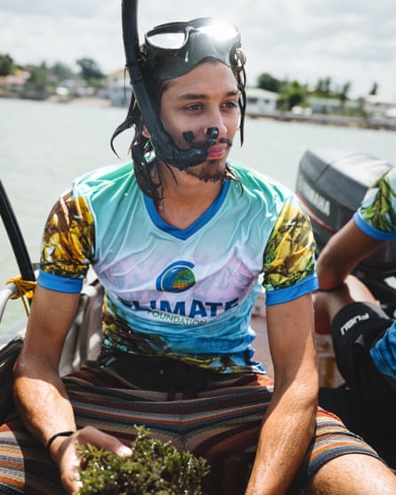 A young man with a mask and snorkel sits on a boat holding a clump of seaweed 