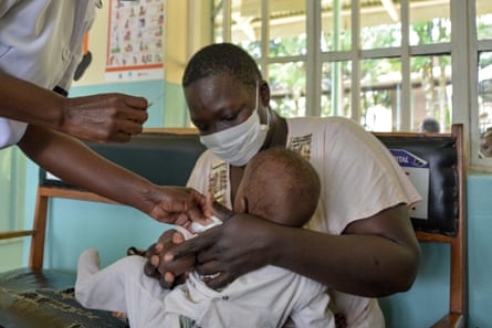 A man in a surgical mask holds a toddler while another person swabs the child’s arm in preparation for a vaccination.