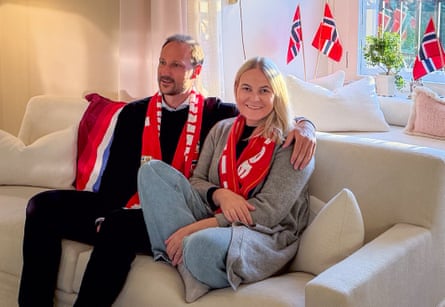 Haakon and Mette-Marit wearing Norway scarves sit on a sofa with Norway flags behind them