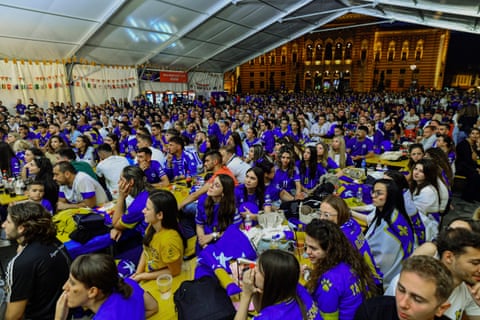 Crowds of people sitting in a large hall, weraing both flags