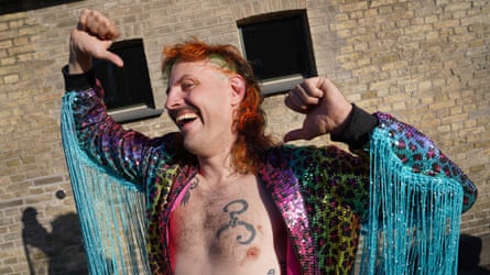 Man with a red mullet and a sparkly outfit shows off his hair and chest tattoo