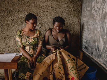 A young woman watches while another sews on a piece of sacking.