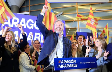 Azcón, standing at a lectern, smiles as he raises a fist into the air, while supporters surround him smiling, cheering and holding the yellow a red striped flag of Aragón